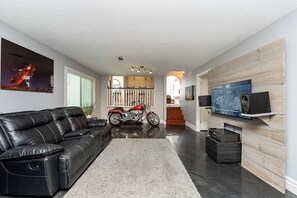A living room with a black leather sofa, a red motorcycle, a flat-screen TV mounted on a wooden wall, and a staircase leading to a kitchen area. - East Abbotsford Spanish Villa (Abbotsford)