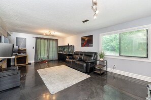 A modern living room with a dark floor, a leather sofa, a white rug, a television, and a window with blinds. - East Abbotsford Spanish Villa (Abbotsford)