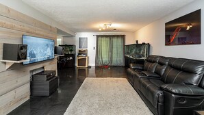 A modern living room with a flat-screen TV, leather sofa, and a large window with curtains. - East Abbotsford Spanish Villa (Abbotsford)