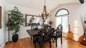 A dining area with a wooden table, dark chairs, a chandelier, a potted plant, a guitar, and a mirror on the wall. - East Abbotsford Spanish Villa (Abbotsford)