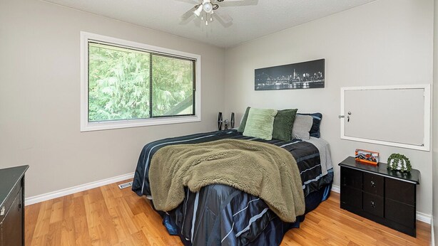 A bedroom with a bed, a ceiling fan, a window with blinds, a cityscape picture, a door, and a small cabinet. - East Abbotsford Spanish Villa (Abbotsford)
