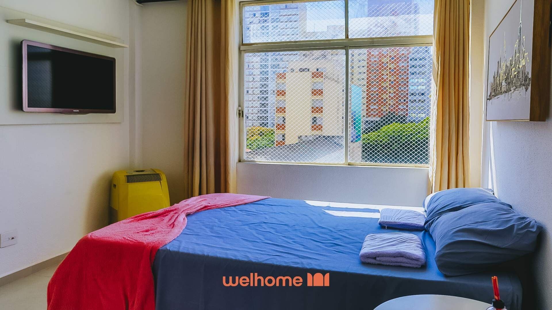 Quarto de hotel bem iluminado com uma cama, uma televisão, uma janela com vista para prédios e uma mala amarela.