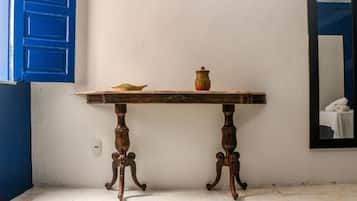 Mesa de madeira com um vaso e uma tigela, posicionada em frente a uma parede branca com uma porta azul e um espelho refletindo uma toalha branca.