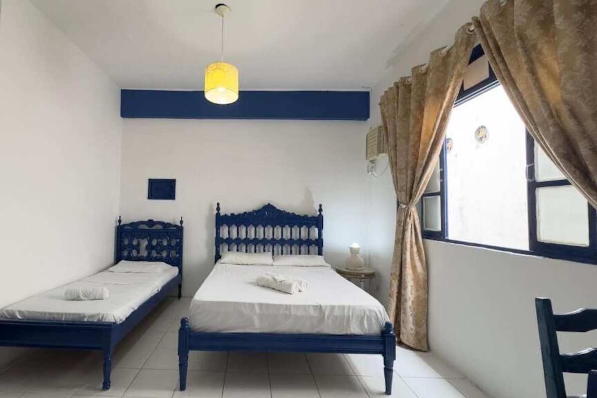 A room with two beds, a bedside table, a hanging light fixture, and a window with curtains.