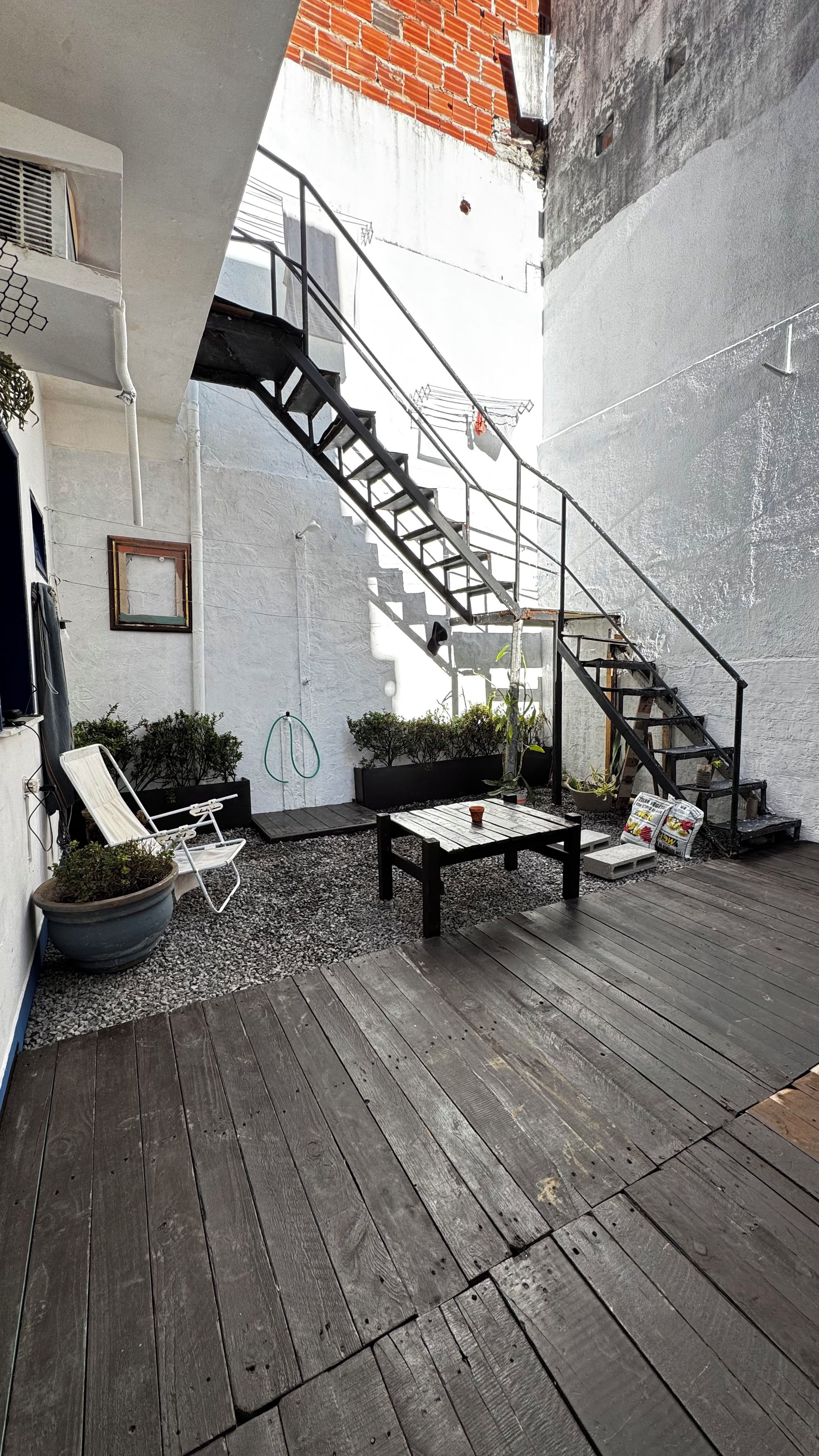 A wooden deck with a metal staircase leading to a white wall with a red brick pattern. There are potted plants and a metal chair.