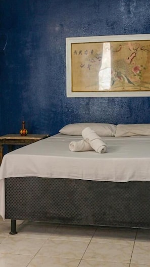 A neatly made bed with a rolled towel, a framed artwork on the wall, and a small table with a lamp.