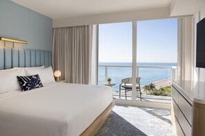 Ein Hotelzimmer mit Bett, Fernseher, einem großen Fenster mit Meerblick und einem Balkon mit Stuhl.