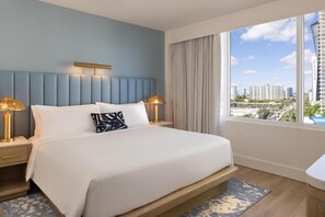 Ein Hotelzimmer mit einem großen Bett, zwei Nachttischlampen, Blick auf die Stadt durch große Fenster und einer sichtbaren Stadtansicht.