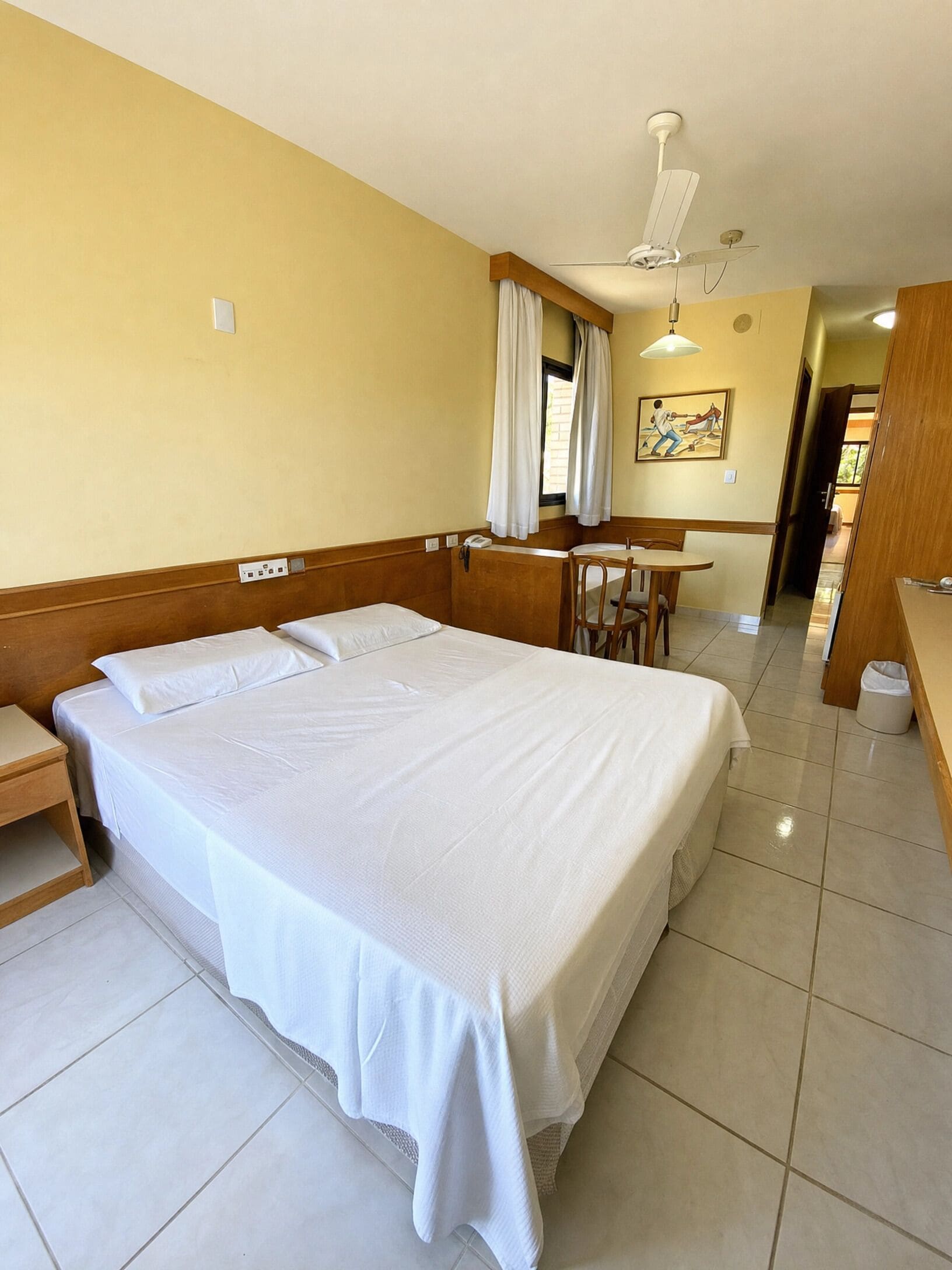 Quarto de hotel com uma cama grande, uma escrivaninha, uma cadeira, um ventilador de teto e um quadro na parede.