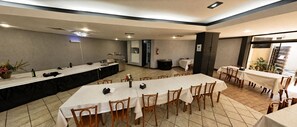 Un salón de conferencias con mesas largas, sillas, una zona de cocina y vistas a un pequeño jardín.