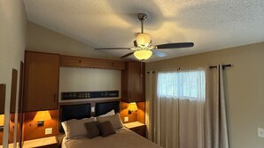 Ein Hotelzimmer mit Bett, Holz-Kopfteil, Nachttischlampen, Deckenventilator und einem Fenster mit Vorhängen.