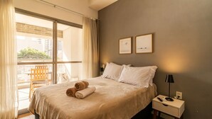 Habitació d'hotel amb un llit gran, dos almoinos, una tauleta de nit amb una làmpada, una cadira i vista a edificis per la finestra.