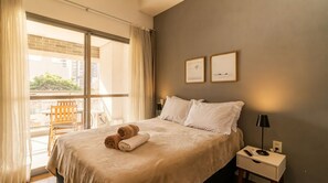 A hotel room with a large bed, two pillows, a nightstand with a lamp, a chair, and a view of buildings through the window. - Studio com Varanda perto do Metrô em Pinheiros (São Paulo)