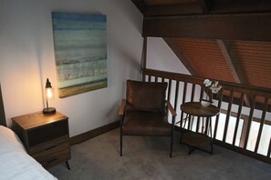 A bedroom with a bed, a nightstand with a lamp, a brown leather chair, a small round table with a flower, and a painting on the wall. - Ski in Style @ Sapphire Squirrel (Wintergreen Resort)