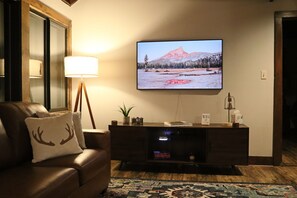 A living room with a leather couch, a flat-screen TV mounted on the wall, a floor lamp, and a decorative rug. - Ski in Style @ Sapphire Squirrel (Wintergreen Resort)