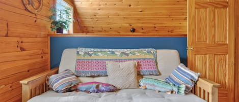 Interior de una cabaña de madera con sofá cama, almohadas, una planta colgante y una puerta.