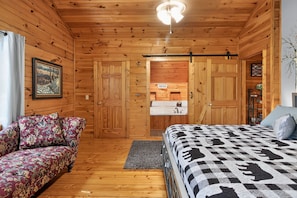 Interior de una cabaña de madera con una cama, un sofá, un baño con lavabo y un cuadro en la pared.