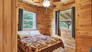 Un dormitorio de cabaña de madera con una cama, dos ventanas y un ventilador de techo.
