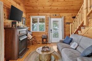 A cozy cabin interior with a fireplace, a television, a sofa, and a staircase. - Scenic 4BR Retreat w/ Balcony & Mountain Views (Hartford)