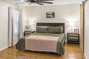 A bedroom with a bed, two bedside tables, a ceiling fan, a window with curtains, and a painting on the wall.