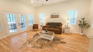 A living room with a brown sofa, a coffee table, and a rug. There are two windows with blinds, a door with glass panels, and a plant.