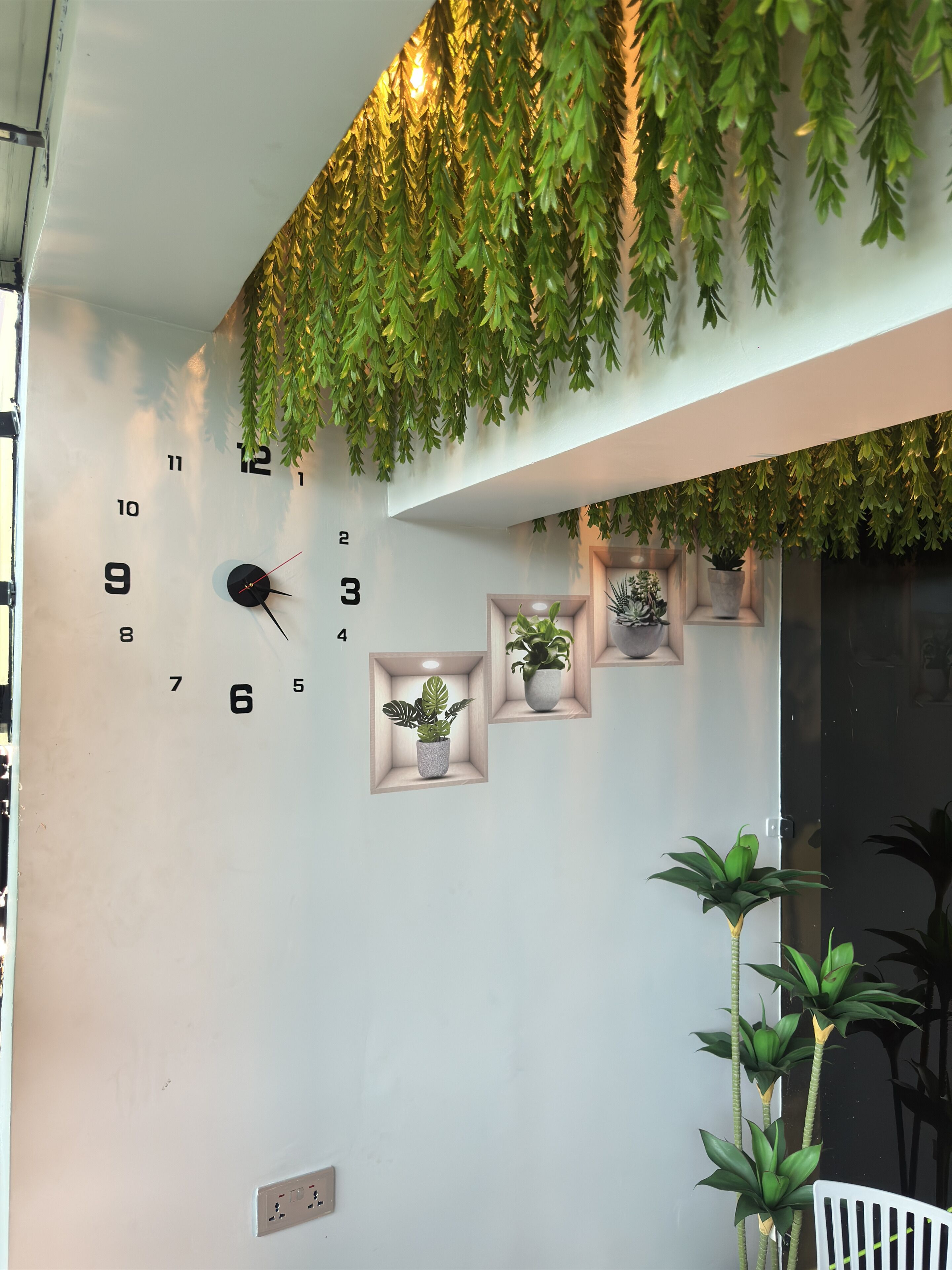A wall with a clock, potted plants, and a hanging green foliage decoration.