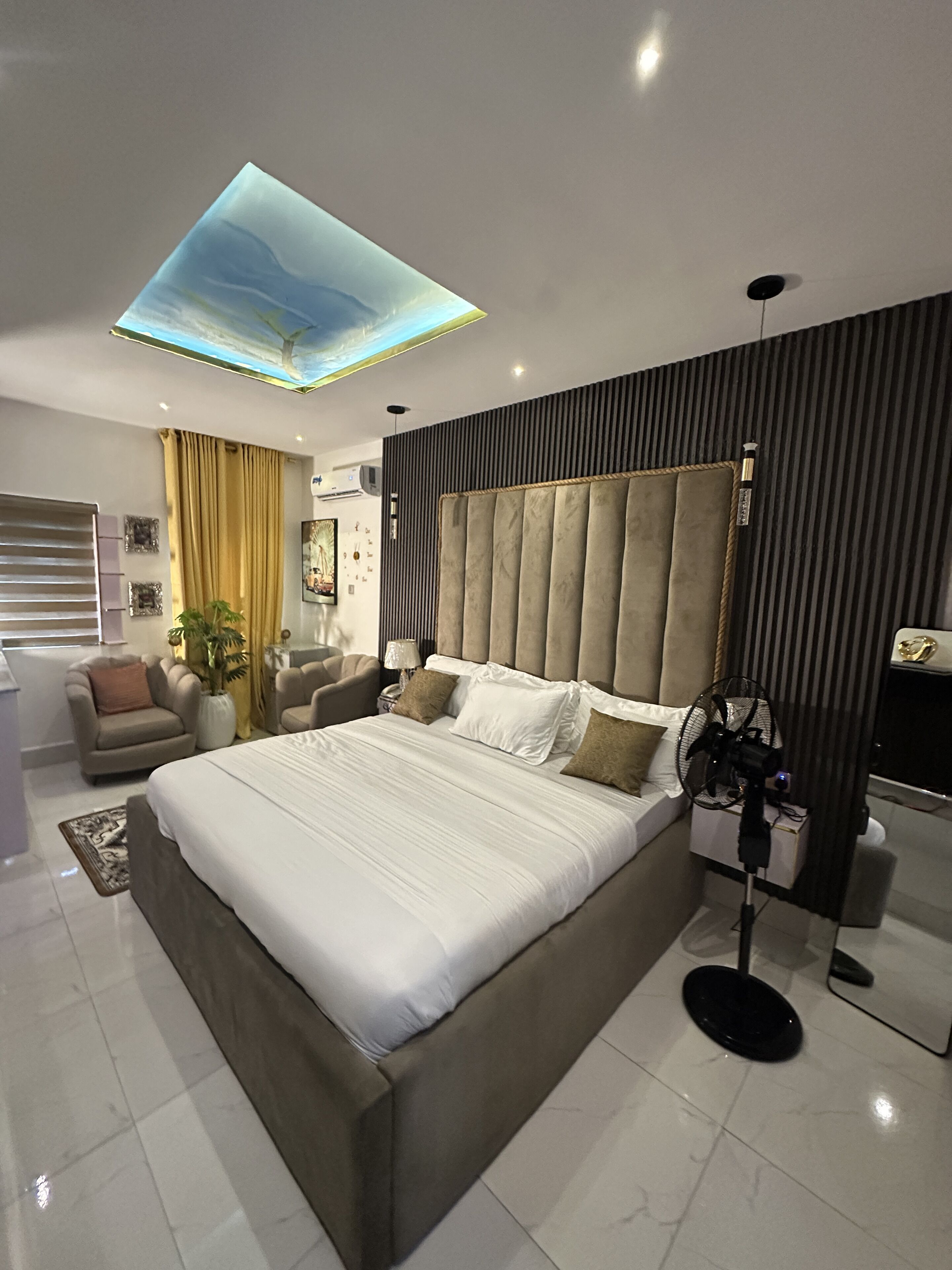 A modern bedroom with a large bed, a ceiling light, a bedside table, a fan, and a window with curtains.