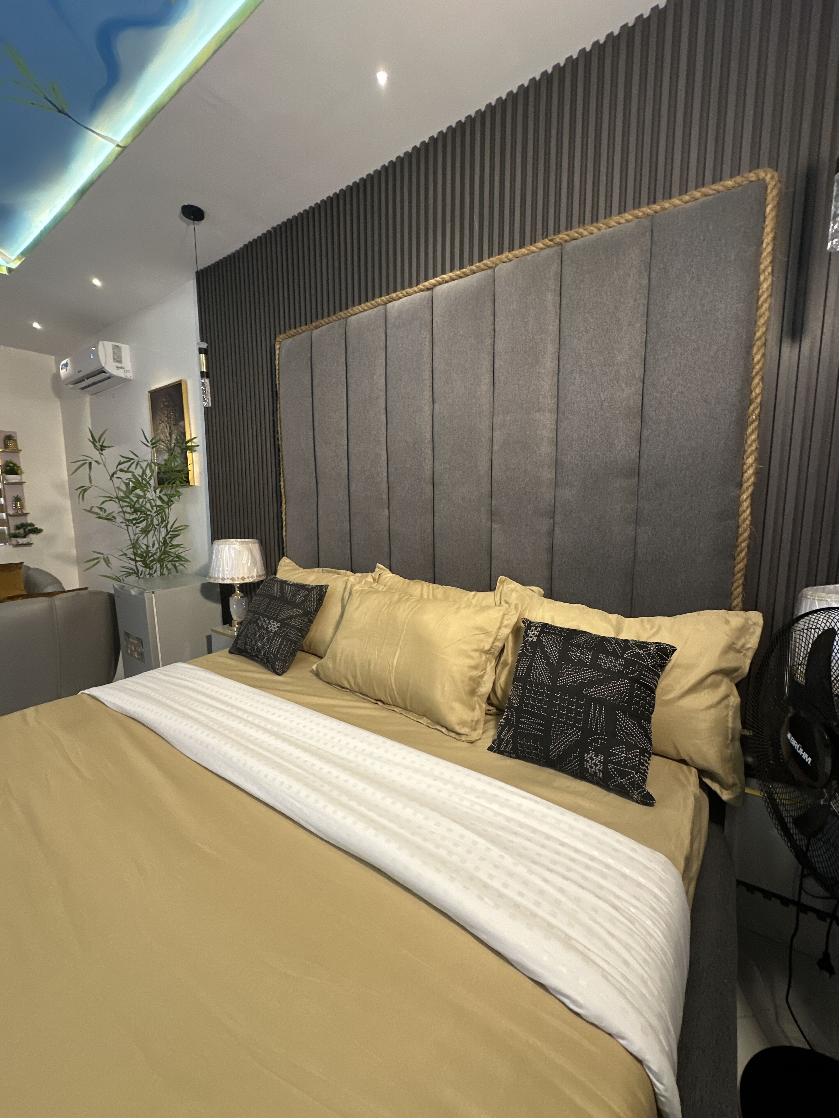 A modern bedroom with a large bed, a headboard with vertical panels, a bedside table with a lamp, and a wall-mounted air conditioner.