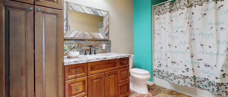A bathroom with wooden cabinets, a marble countertop, a white toilet, and a teal shower curtain with motivational text.