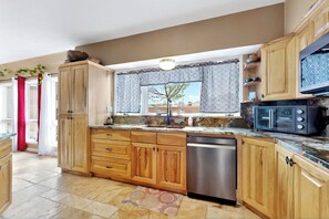 A kitchen with wooden cabinets, a stainless steel dishwasher, a microwave, and a window with patterned curtains. - Entire Desert Hacienda Estate Compound - Sleeps 12 in La Mesilla, New Mexico (Española)