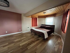 Een slaapkamer met houten vloer, een bed, een raam met gordijnen en een plafondventilator.