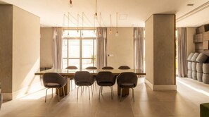 A modern dining area with a long table and six chairs, large windows with curtains, and hanging pendant lights. - Studio com Varanda Perto do Metr em Pinheiros (São Paulo)
