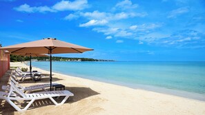 A beach with white lounge chairs and a large umbrella, a clear blue sky, and calm sea. - Spacious Two-Bedroom Kitchenette within a Beachfront Property in Casual Negril (Negril)