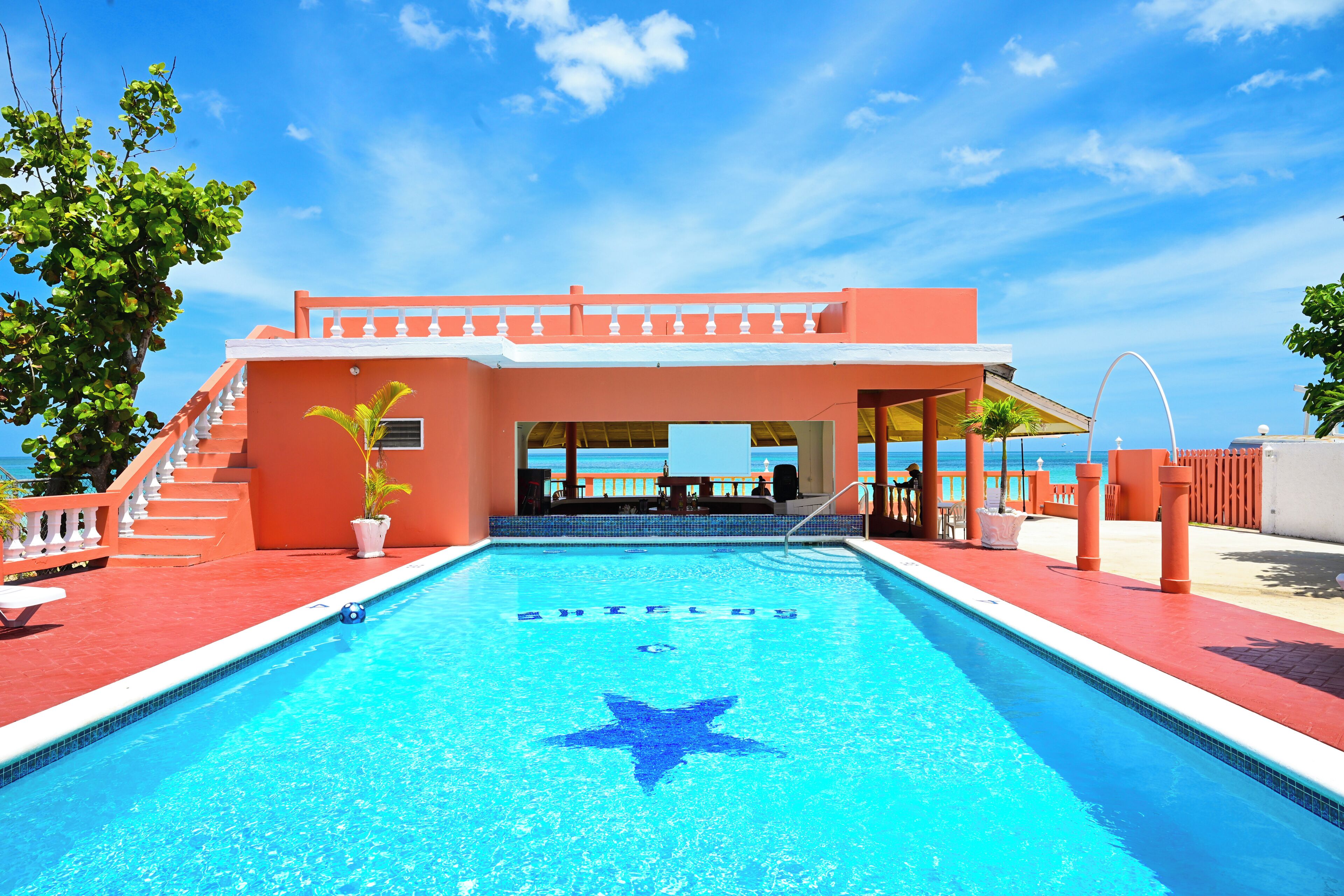Une piscine en forme d’étoile, entourée d’un bâtiment rouge doté d’un balcon blanc, et offrant une vue sur l’océan.