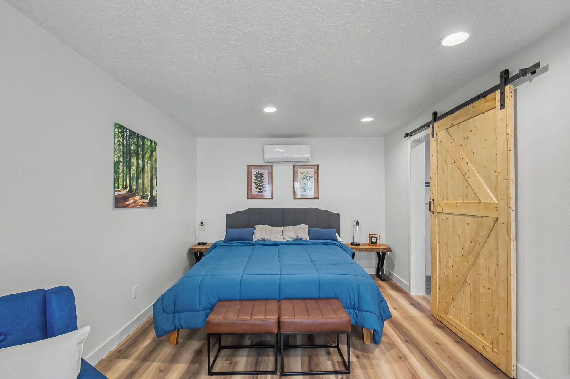A bedroom with a bed, a bench, a wooden door, and two wall-mounted air conditioners.
