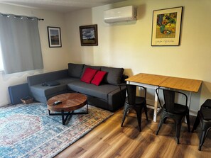 A living room with a grey sofa, a small round table, a wooden dining table with two chairs, a patterned rug, and two framed pictures on the wall. - Cute clean cottage with modern amenities near downtown (McMinnville)