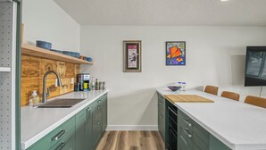 A modern kitchen with green cabinets, a white countertop, and a wooden backsplash. There is a coffee maker, a sink, and a dining area with a table and chairs. - Cute clean cottage with modern amenities near downtown (McMinnville)