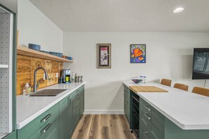 A modern kitchen with green cabinets, a white countertop, and a wooden backsplash. There is a coffee maker, a sink, and a dining area with a table and chairs. - Cute clean cottage with modern amenities near downtown (McMinnville)