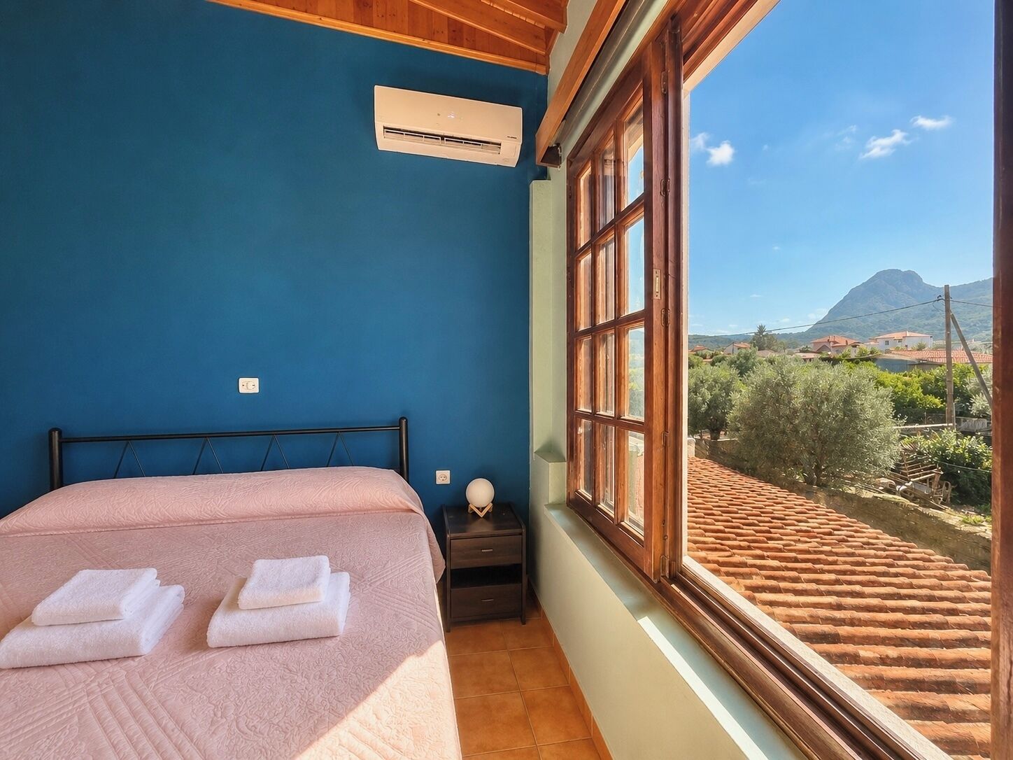 Un dormitorio con una cama, un equipo de aire acondicionado, una mesita de noche y una ventana con vistas a la montaña y casas.
