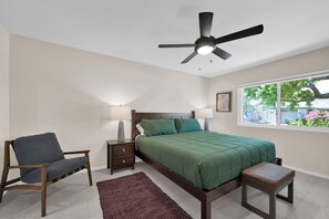 Una camera da letto con un letto, una sedia, un comodino, una lampada, una lampada da terra, una finestra e una ventola a soffitto.
