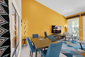 A modern dining area with a glass table, blue upholstered chairs, a flat-screen TV, and a patterned rug. - Nacho's Place! (Palm Springs)