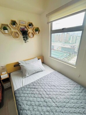 A single bed with a patterned bedspread, a bedside table with a lamp, and a window with a city view. - Cozy Stay at Urban Deca Towers Cubao (Quezon City)
