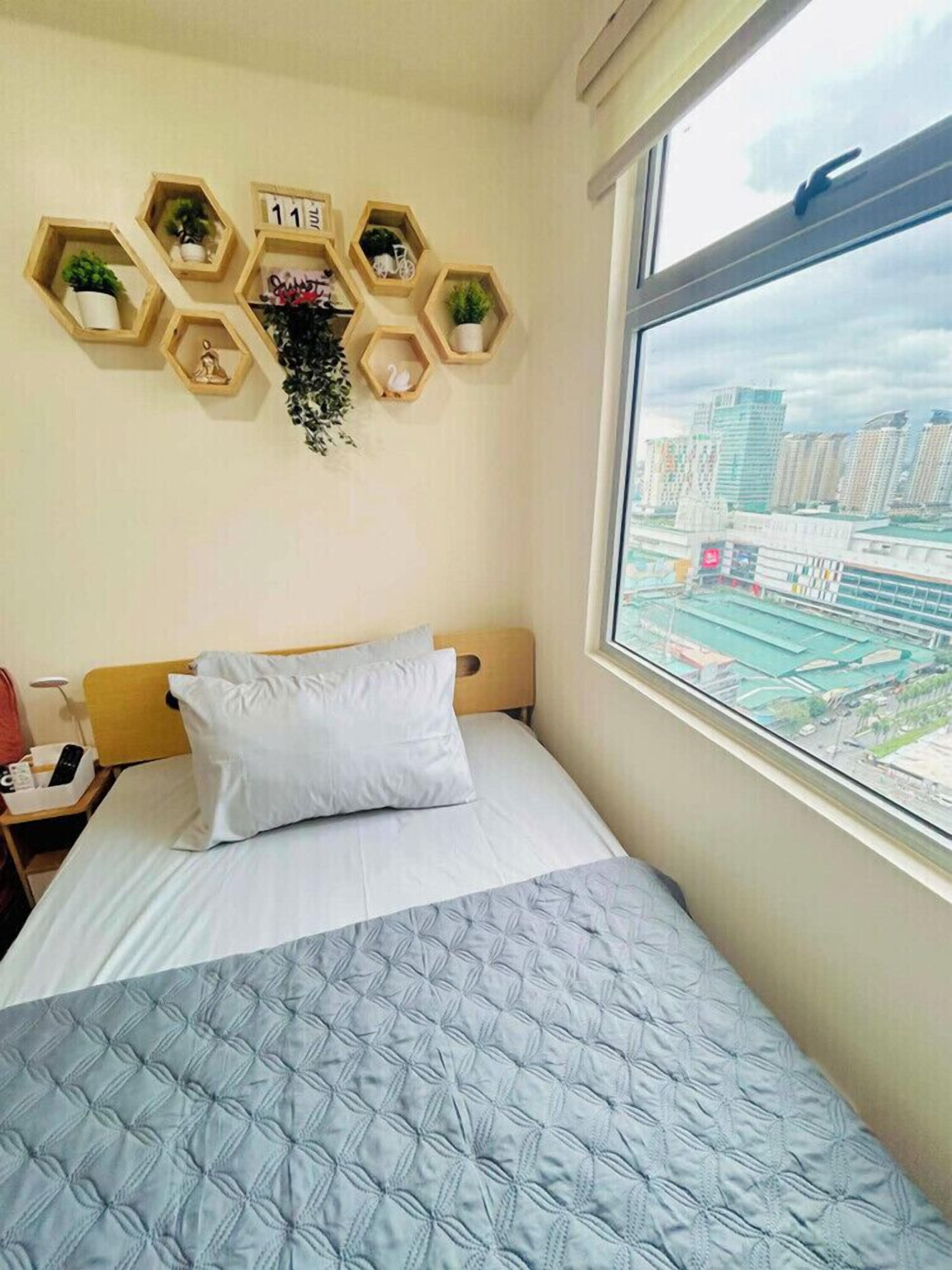 A hotel room with a bed, a window with a city view, and a wall-mounted shelf with plants and a clock.