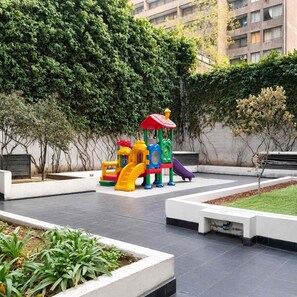 A playground with a slide and climbing structures, surrounded by a landscaped garden and a building in the background. - Family 2BR 2 Baths, Walk to La Moneda-Free Parking (Santiago)