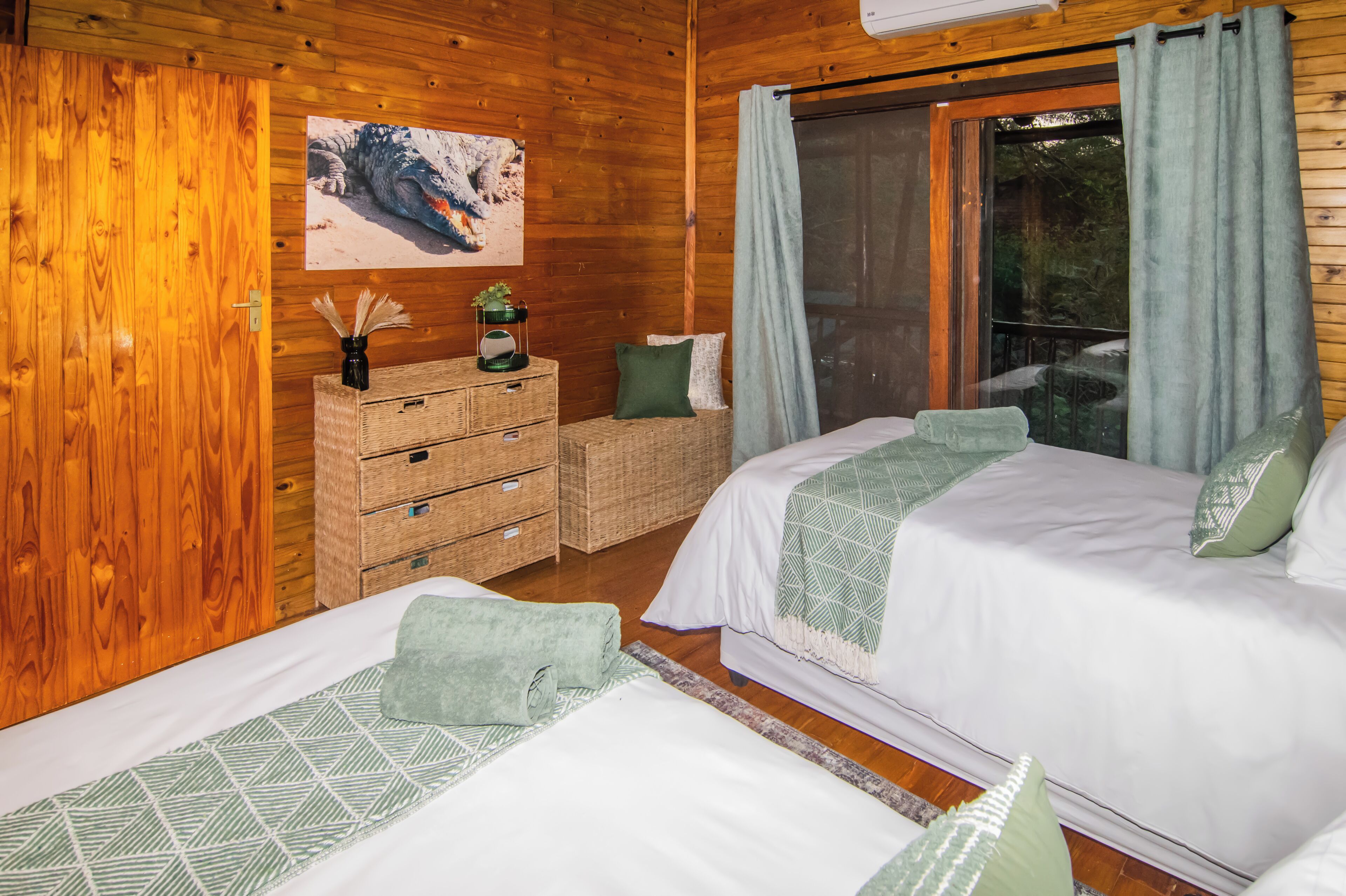 A wooden cabin bedroom with two beds, a dresser, a mirror, and a window with curtains.