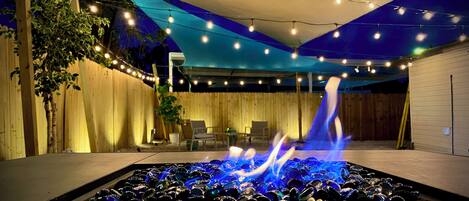 Un patio exterior con una chimenea, luces colgantes y una zona de estar cubierta.