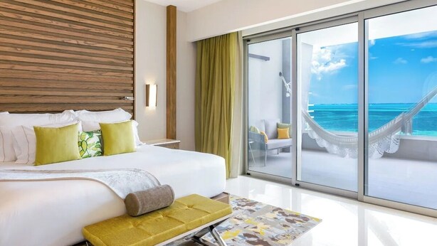 Une chambre d’hôtel avec un grand lit, un banc et une porte vitrée donnant sur un balcon avec vue sur l’océan.
