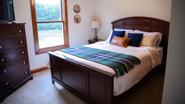 Une chambre à coucher avec un lit en bois, une commode, une fenêtre donnant sur un paysage verdoyant et une lampe sur une table de chevet.