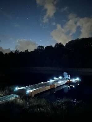 Un muelle que se adentra en el agua, con una silla y una mesita, rodeado de césped y árboles.