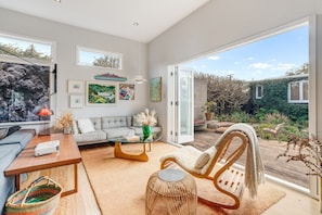A modern living room with a sofa, coffee table, and a chair, a projector, and a view of a garden through large windows. - Pool + Fire Pit + Projector | Walk to Eden Park  | Designer Retreat (Auckland)
